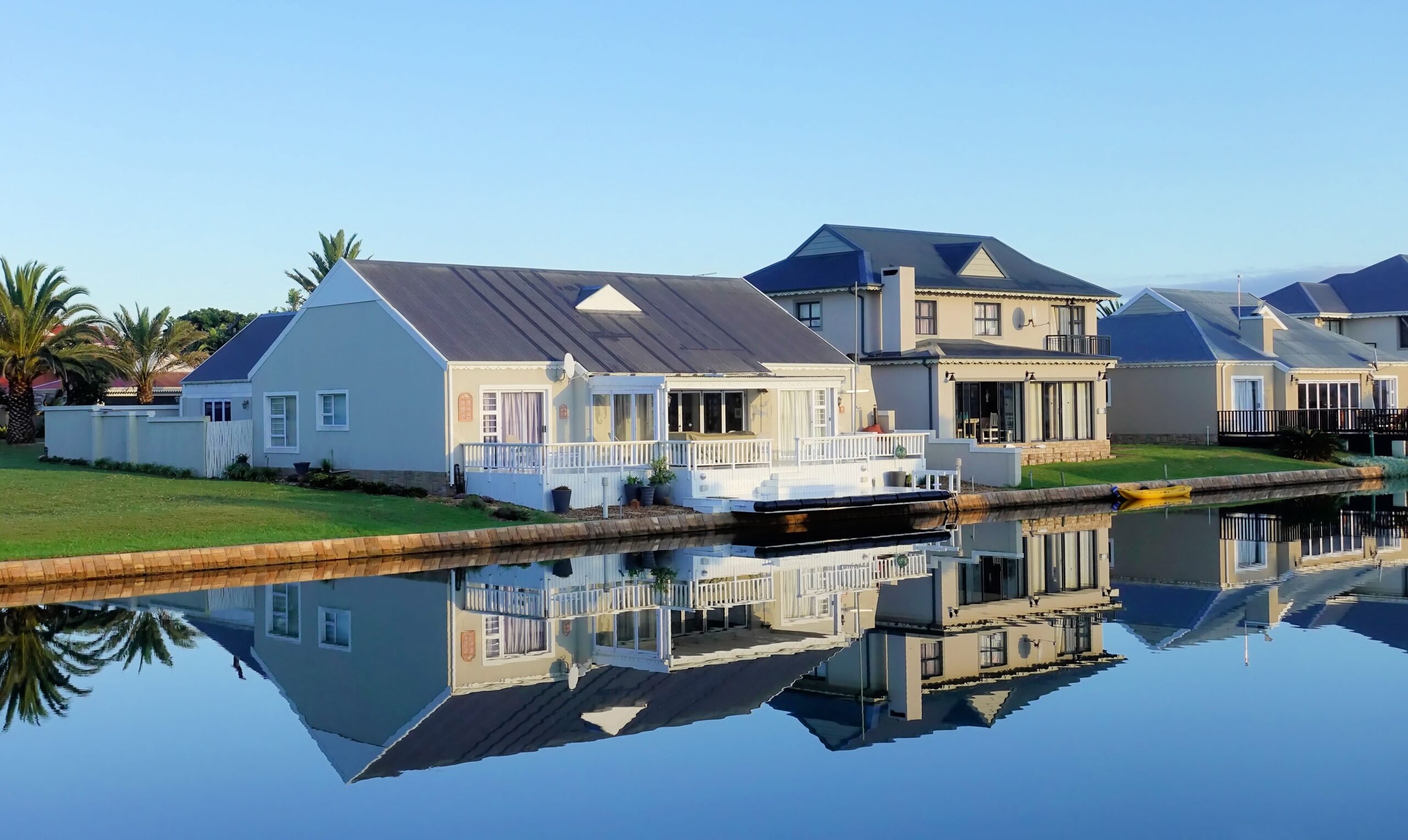 Luxury modern homes reflecting on a peaceful, clear lake during daylight, showcasing architectural beauty.